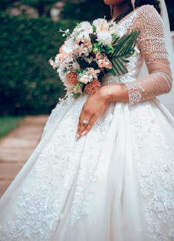 A bride in a stunning wedding dress holds a bouquet, showcasing intricate lace and beadwork details.