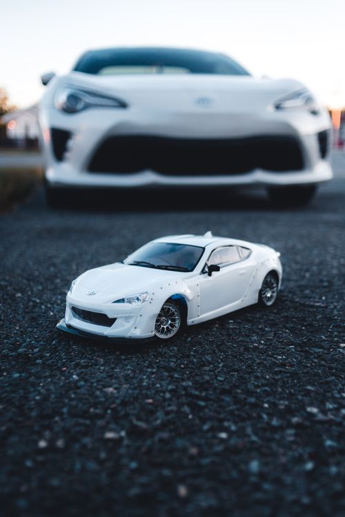 Free A toy sports car placed on the ground with a real car in the background, showcasing automotive contrast. Stock Photo