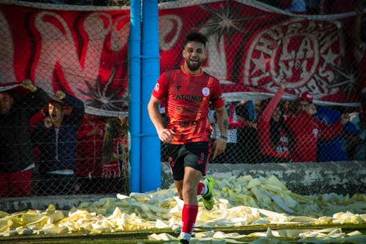 A soccer player in a red jersey celebrates a goal with cheering fans behind during a day match.