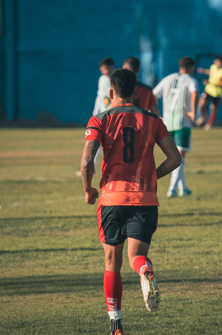 A Person Wearing A Red Jersey Shirt Running In A Grass Field Near People