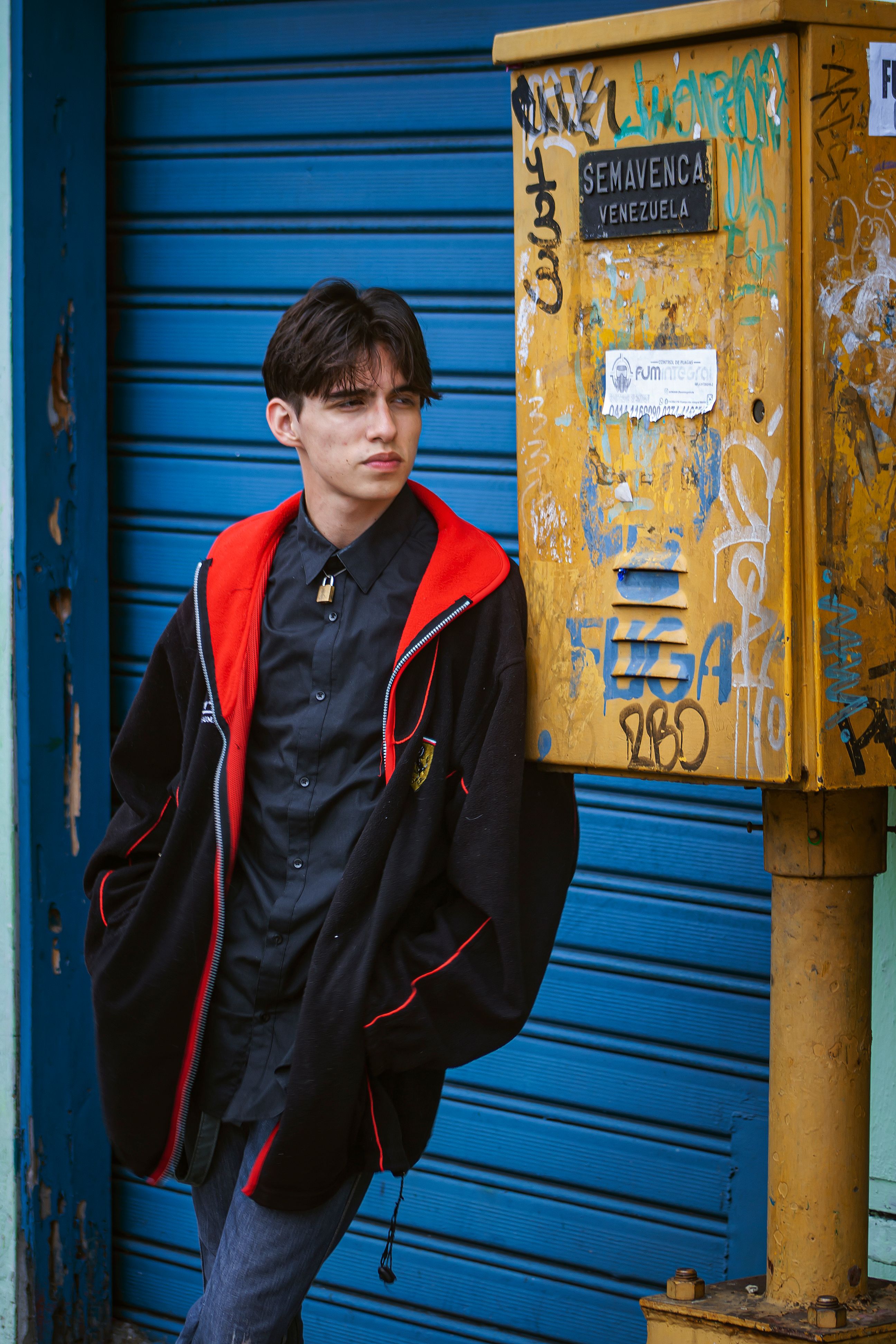 A young man in a black jacket leans against a graffiti-covered box with Venezuelan signage.