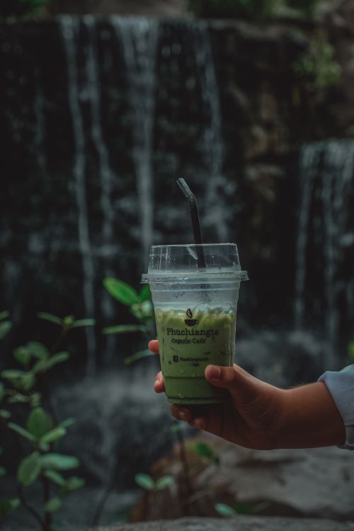 Free Close-up of a green matcha drink held near a scenic waterfall. Stock Photo