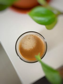 A close-up of a foamy coffee in a glass cup surrounded by green leaves.
