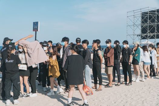 A large group of people wearing masks stand in line at a beach event entrance under clear skies.