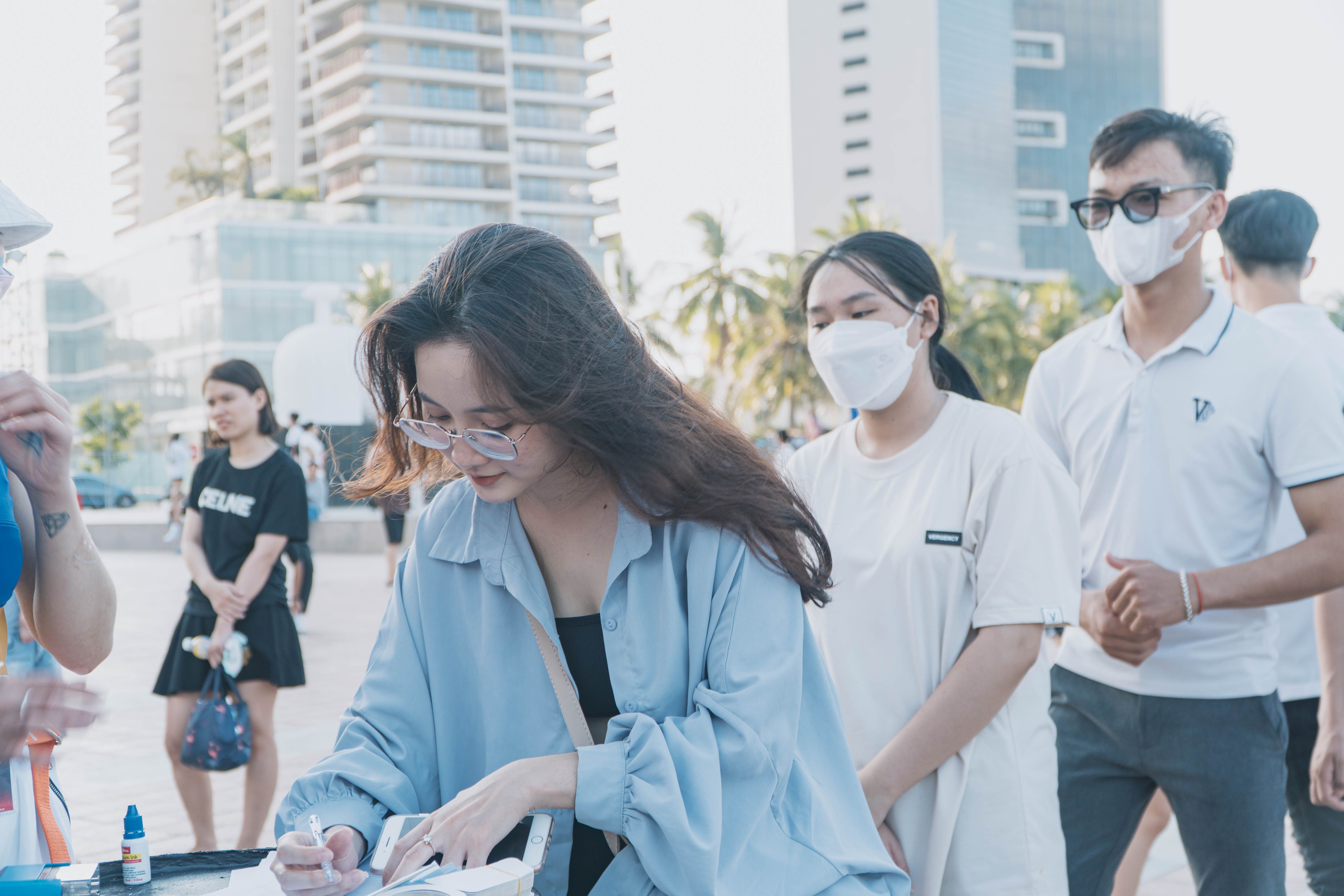 People wearing face masks gather outdoors, focusing on a woman writing. Modern buildings in the background.