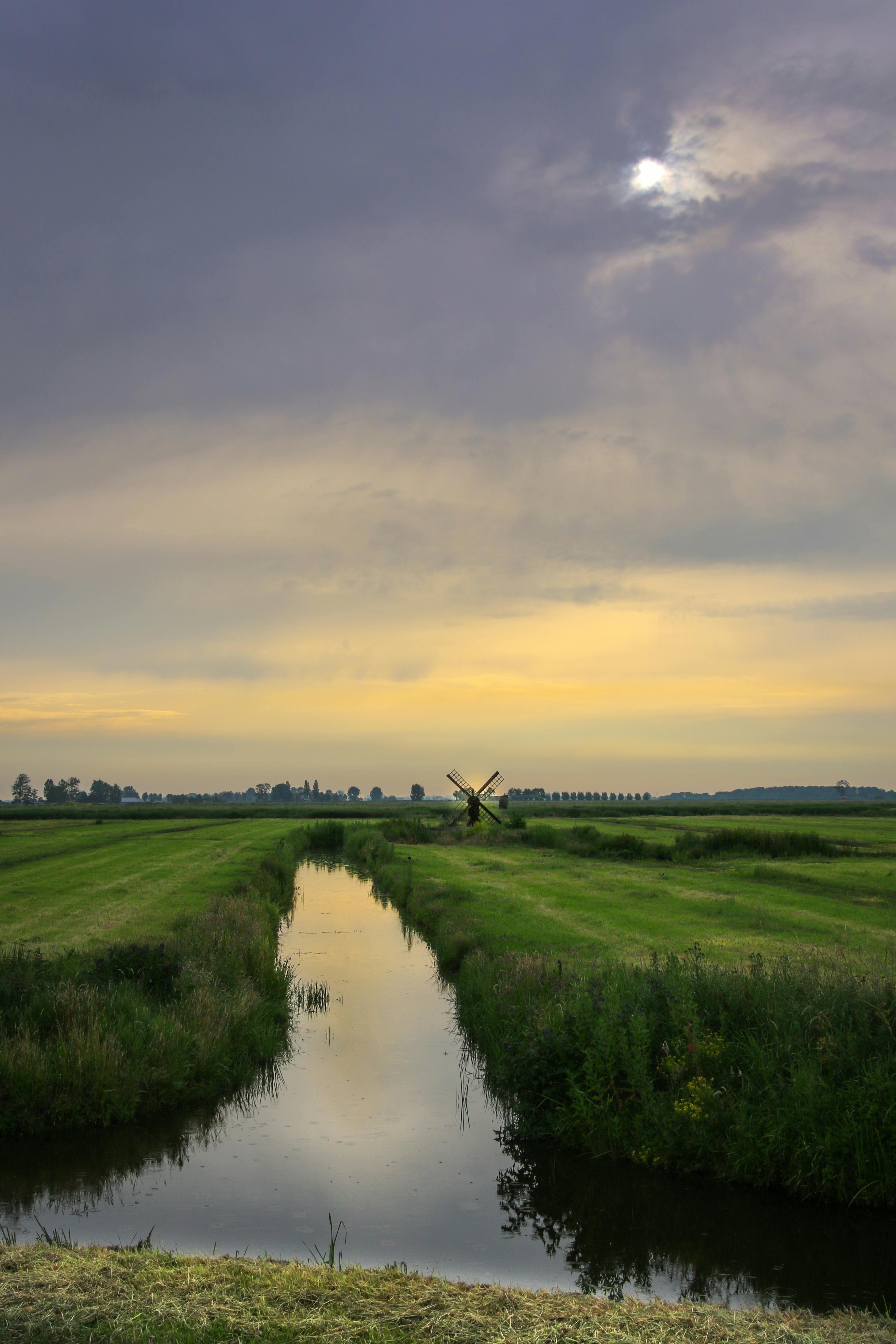 宁静的乡村景观中，河流蜿蜒流淌，田野郁郁葱葱，远处的风车在多云的天空下静静伫立