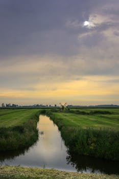 宁静的乡村景观中，河流蜿蜒流淌，田野郁郁葱葱，远处的风车在多云的天空下静静伫立