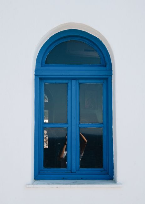 A vibrant blue window set in a classic white wall features a glimpse into Greek architecture.