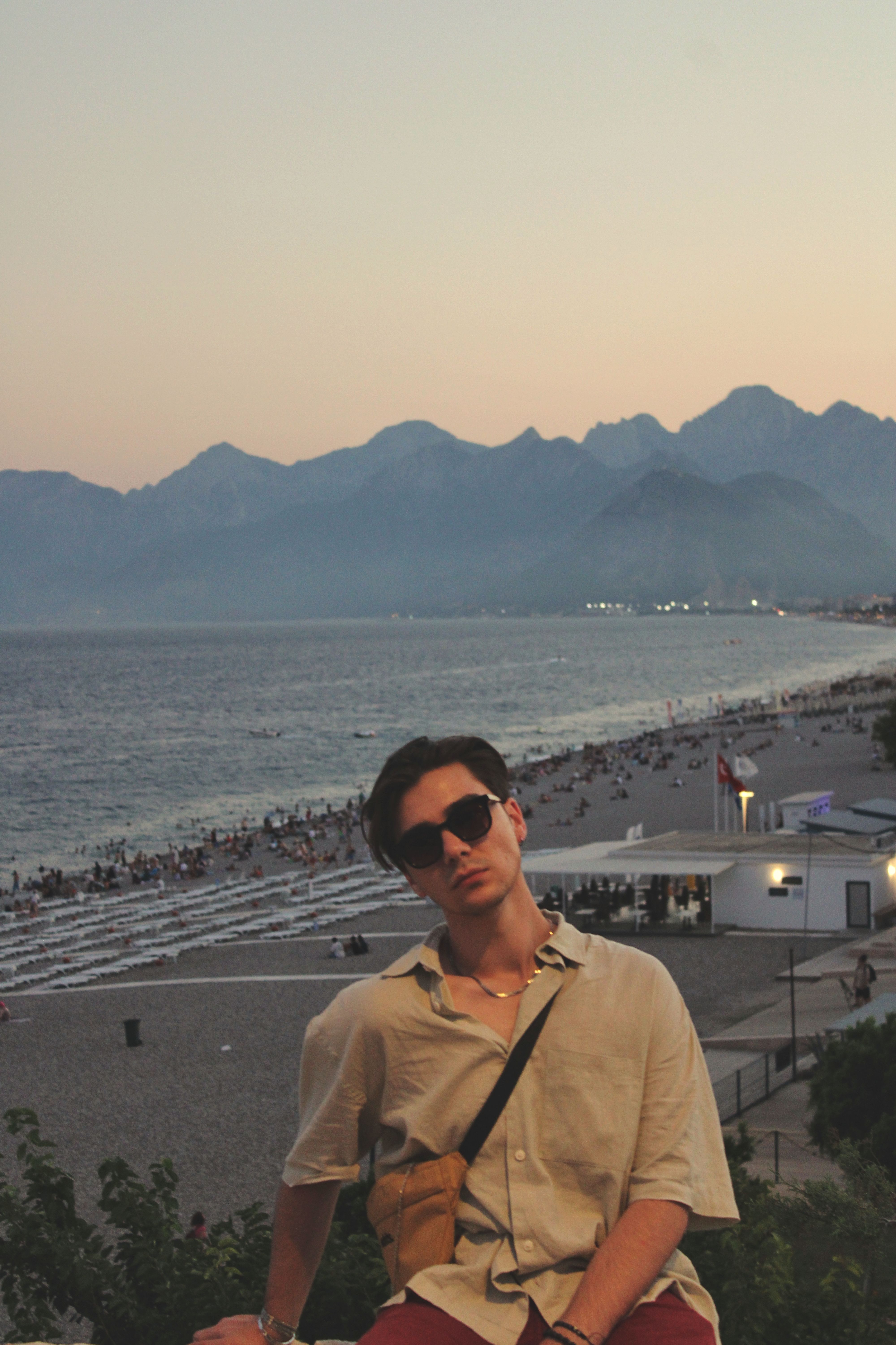 A young man poses at sunset by the beach in Antalya, Turkey, with mountains in the background.