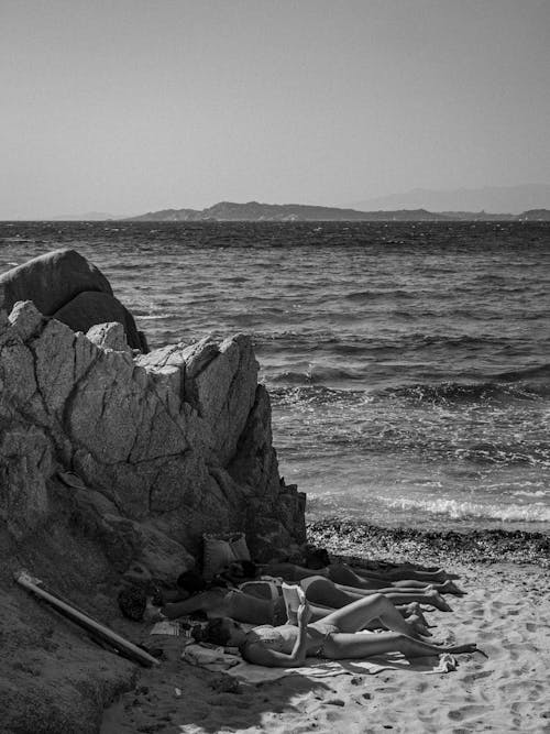 Free Grayscale Photo of People Lying on Sand Beside a Rock Formation on Seashore Stock Photo
