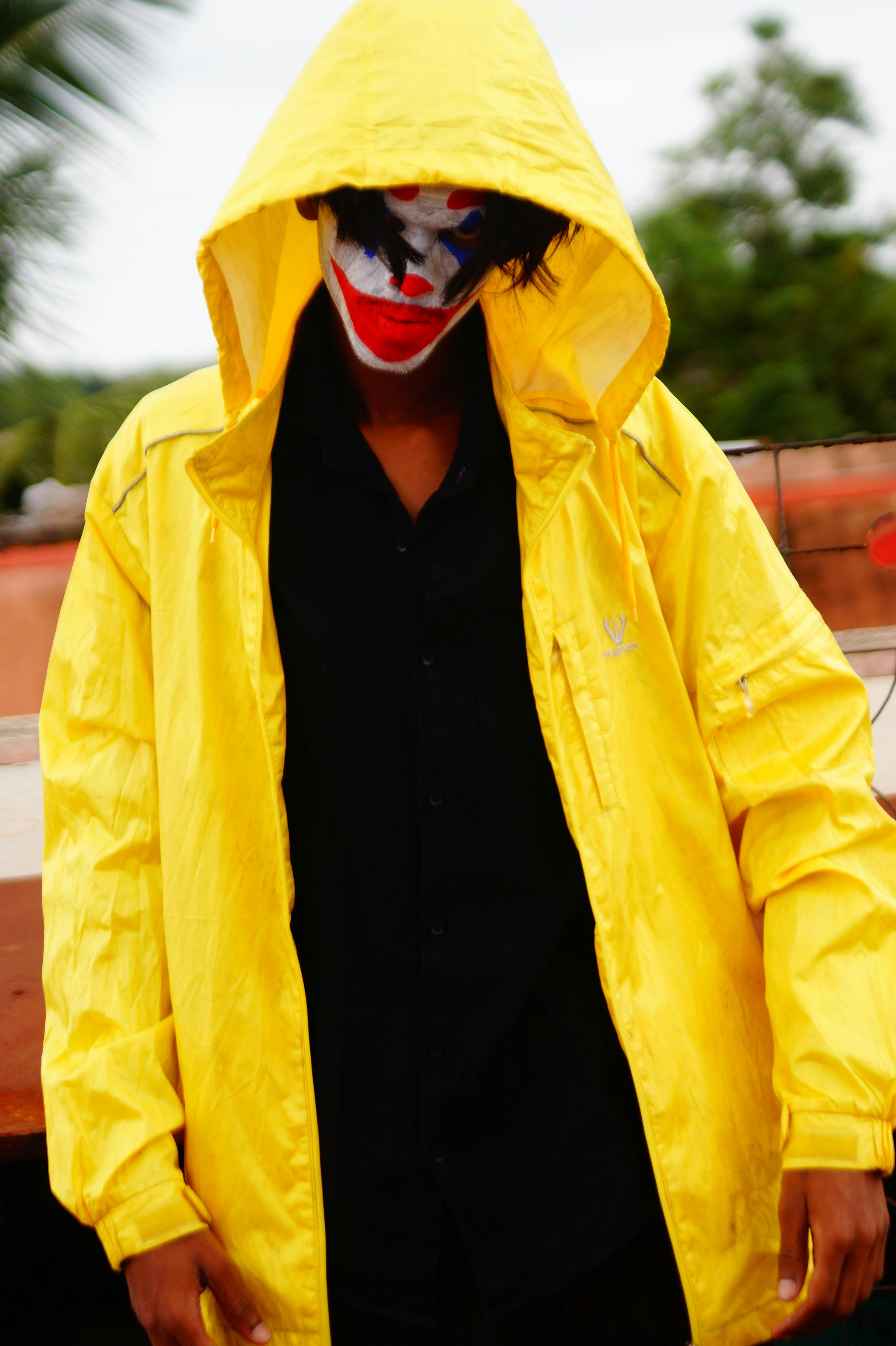 Free A person in a yellow raincoat with joker face paint creates a dramatic and mysterious scene outdoors. Stock Photo