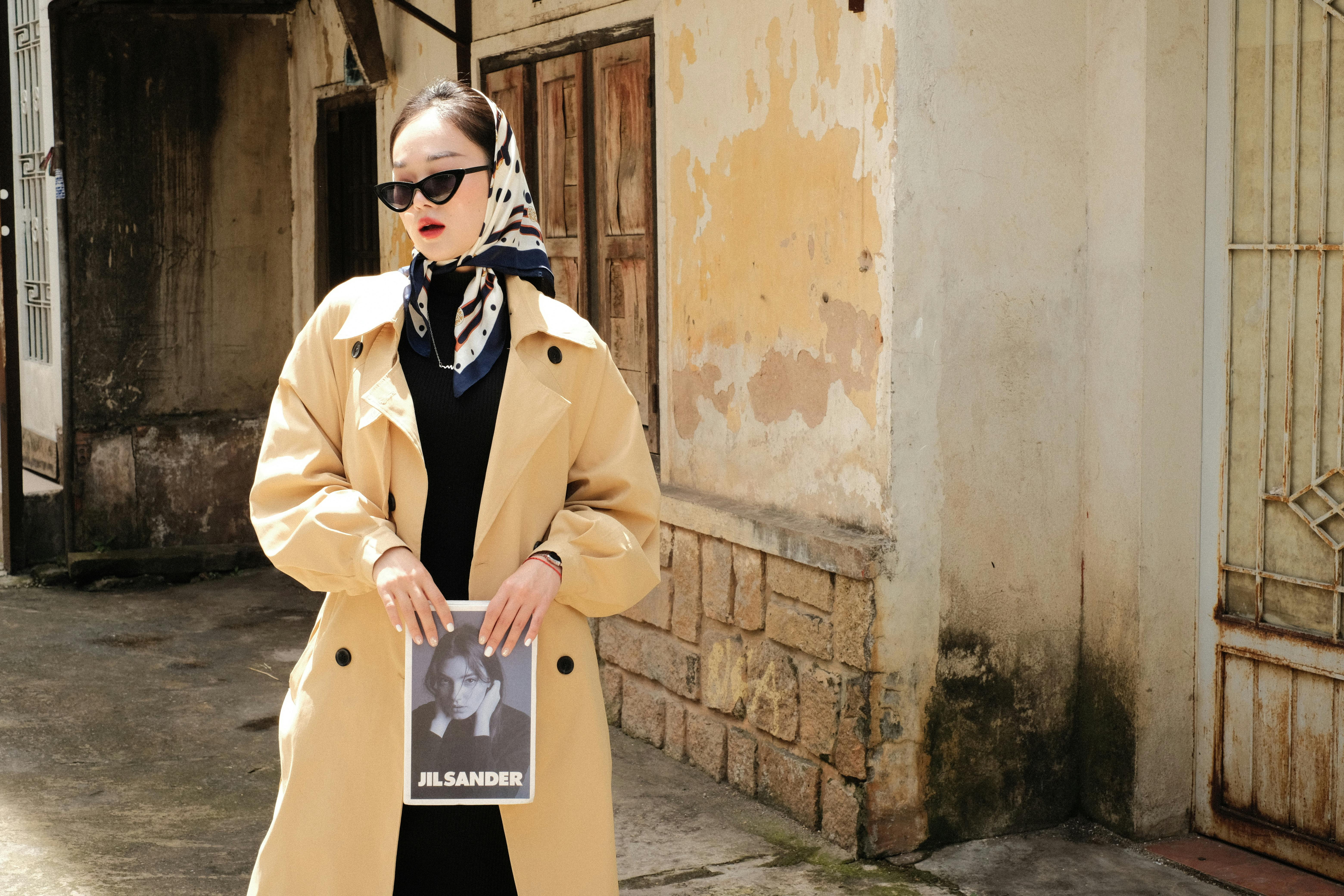 Woman in fashionable trench coat and headscarf holding magazine, posing on a rustic street.