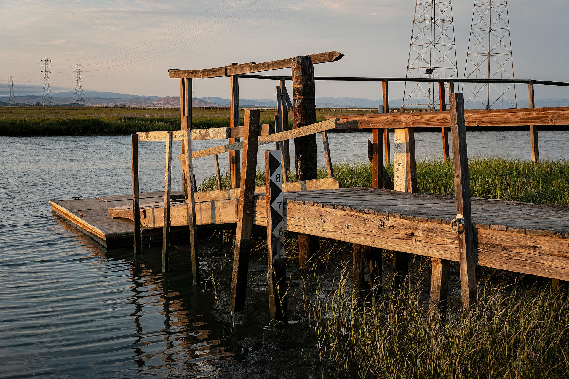 Old wooden dock by a river during sunset with distant hills and towers in view.