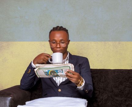 A man in a blue blazer reads a newspaper while drinking coffee indoors.