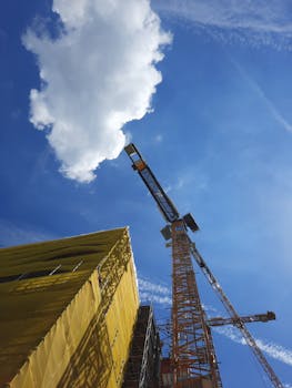 Low angle view of a construction crane and building against a bright blue sky with clouds.