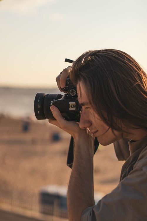Free A thoughtful photographer with long hair takes pictures by the beach during golden hour. Stock Photo
