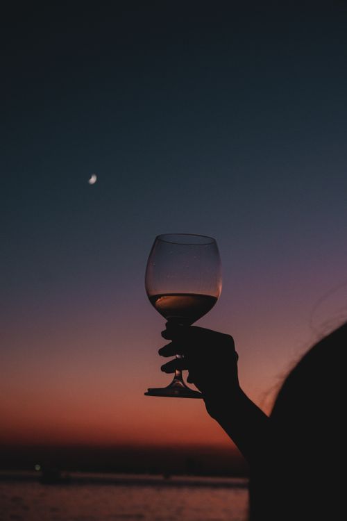 Free A silhouetted hand holding a wine glass against a colorful twilight sky with a crescent moon. Stock Photo