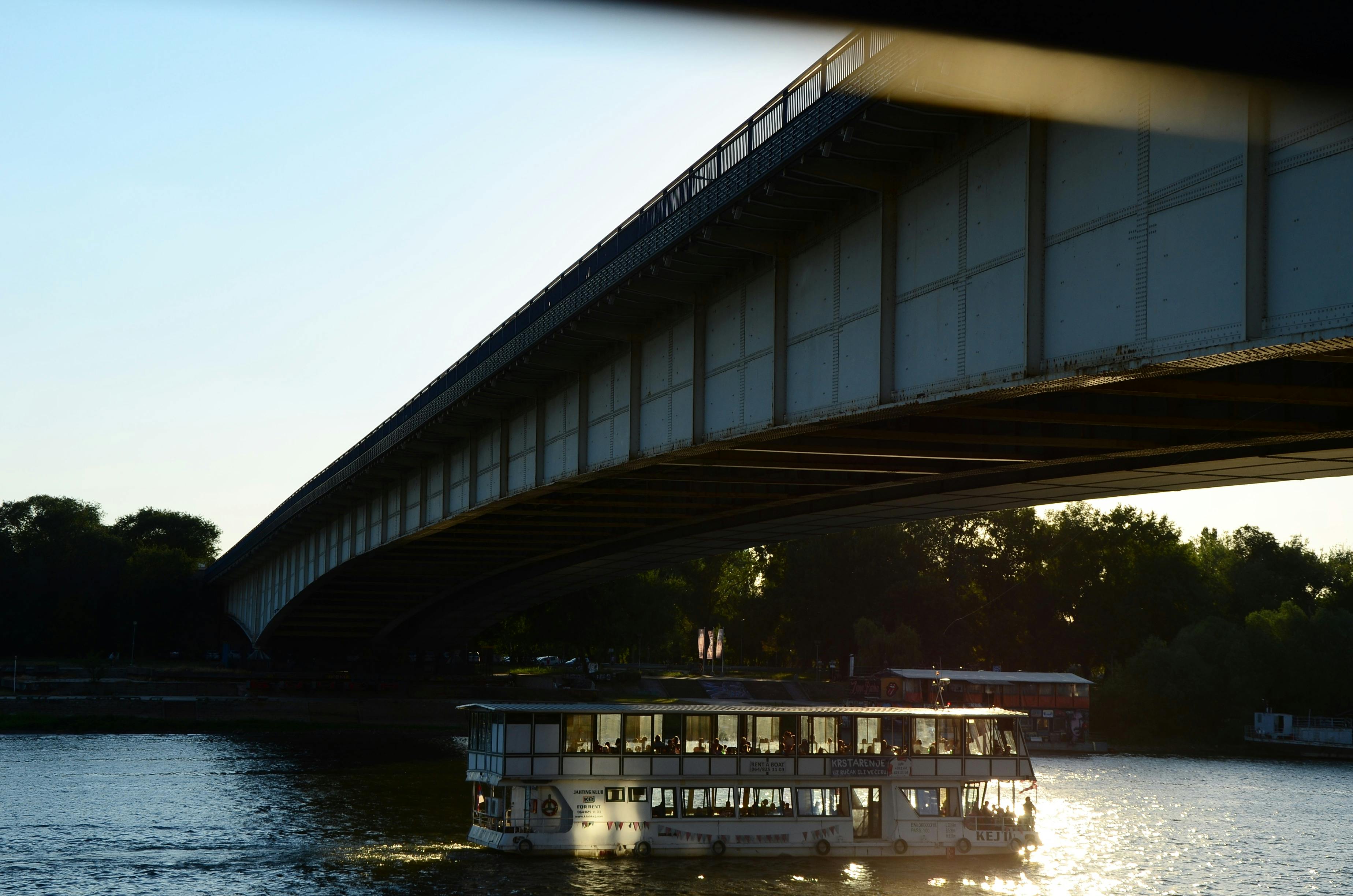 Free A ferry boat gliding under a steel bridge on a sunny day, casting reflections on the river. Stock Photo