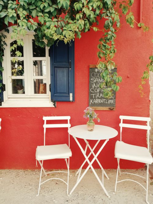 Free Cozy outdoor cafe scene with white chairs and a table against a vibrant red wall and green foliage. Stock Photo