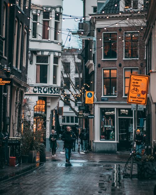 Free A lively Amsterdam street scene featuring hanging lights, cyclists, and wet cobblestones, set in a picturesque urban environment. Stock Photo