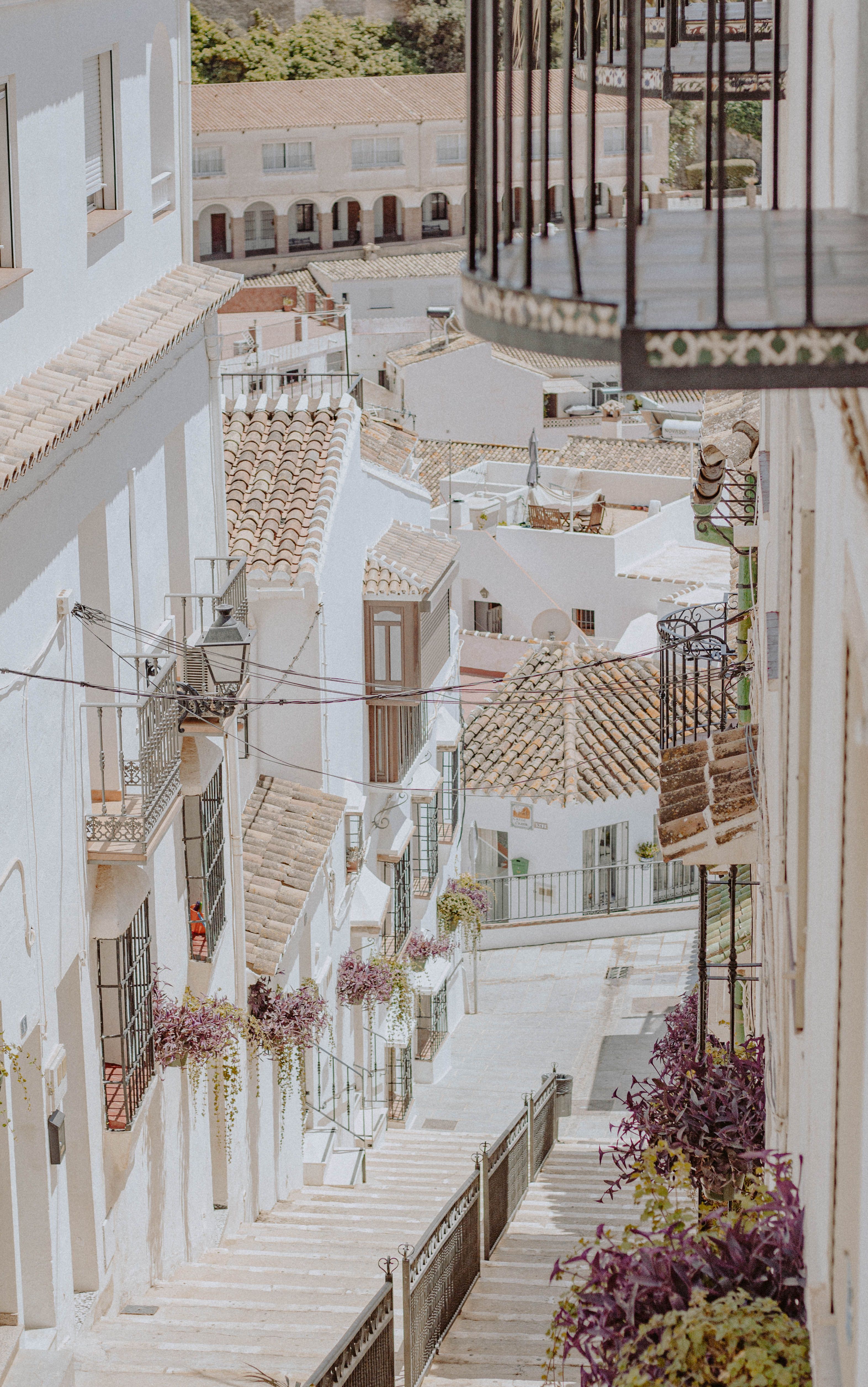 Sunny Mediterranean alley with white walls, rooftops, and hanging flowers.