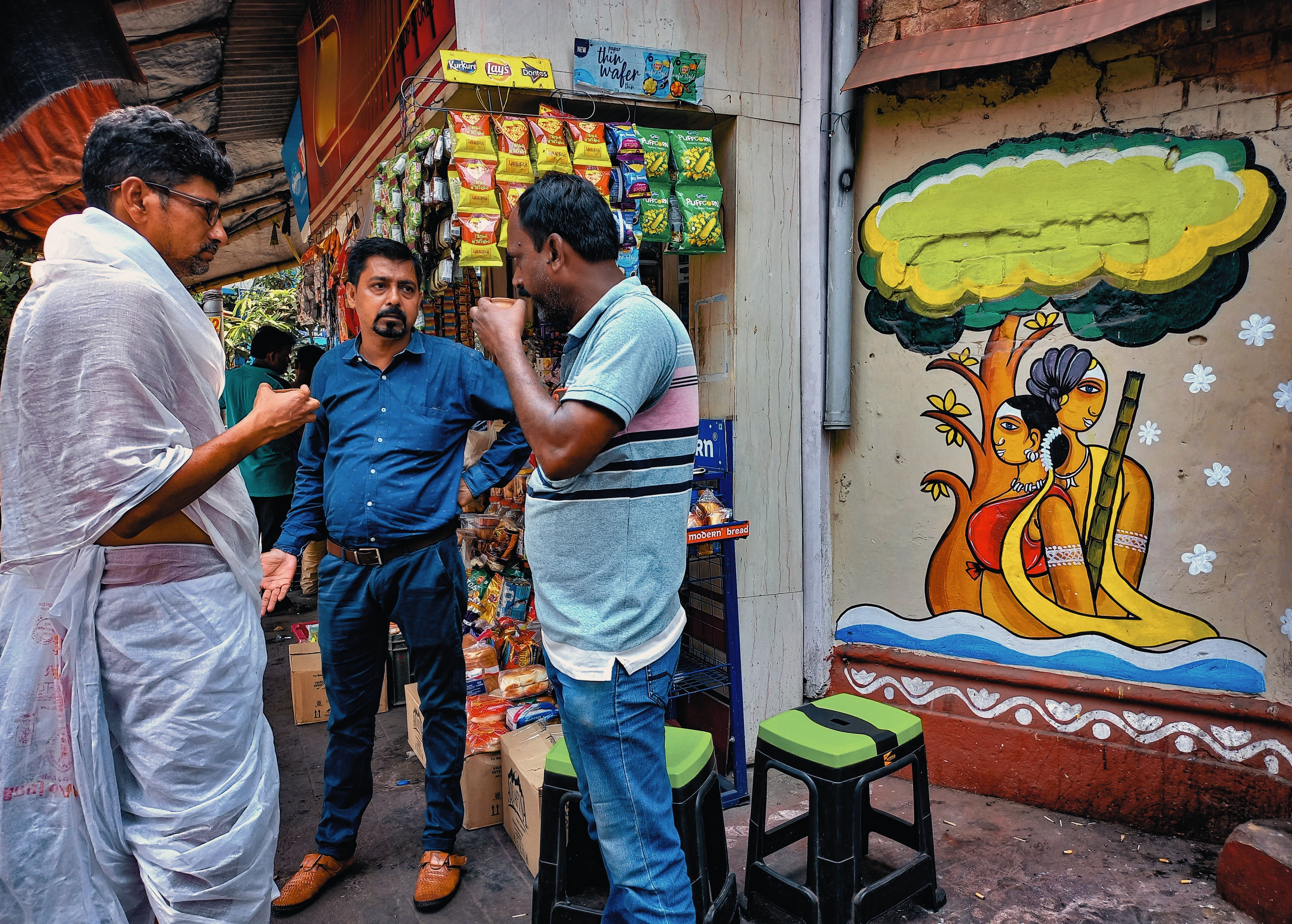 Three men engaging in conversation beside a shop in Kolkata, featuring traditional Indian wall art.