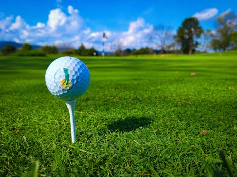 Close-up of a golf ball on a tee against a vibrant golf course with blue skies, perfect for sports enthusiasts.
