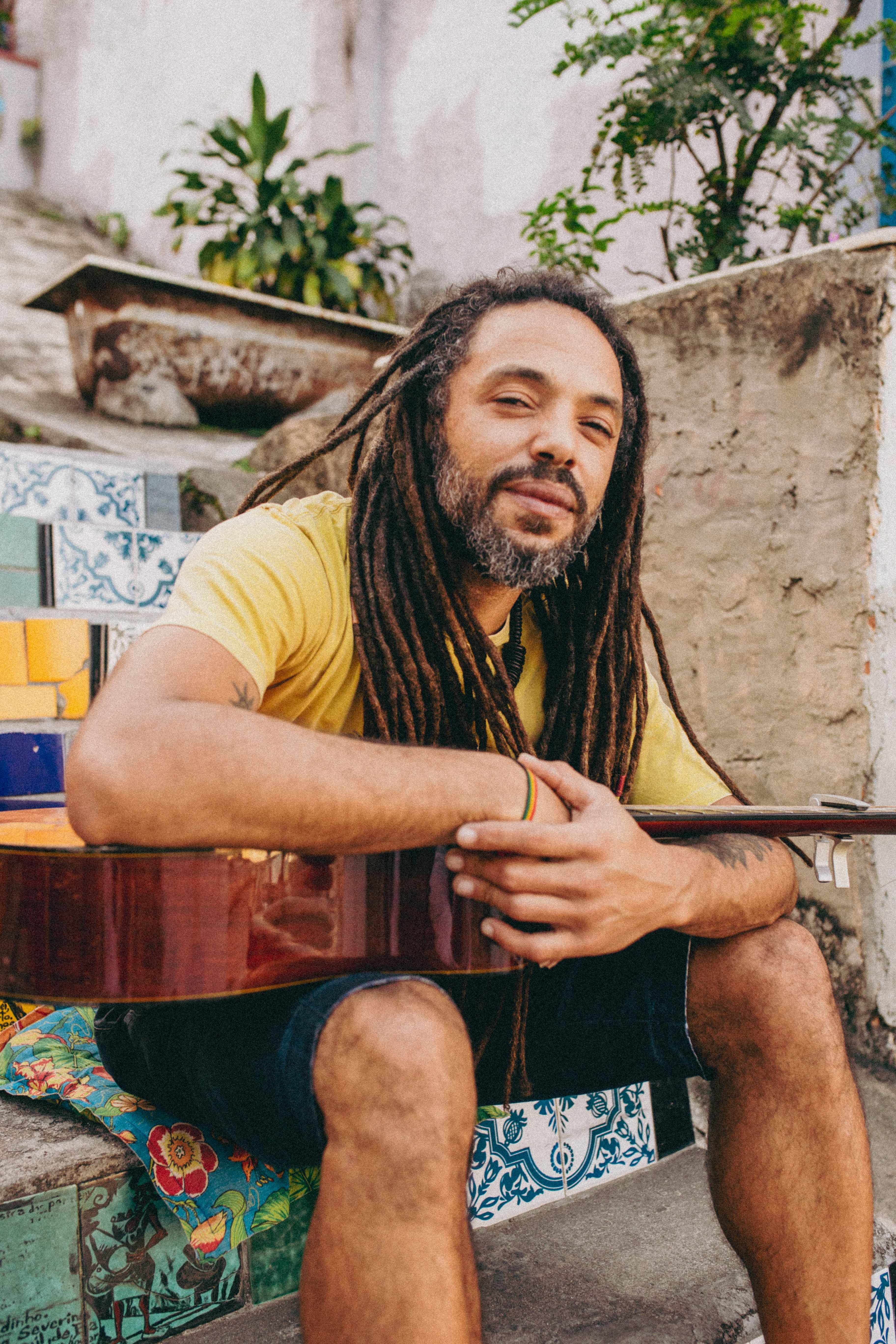 Casual portrait of a dreadlocked musician sitting with a guitar, wearing a yellow shirt.