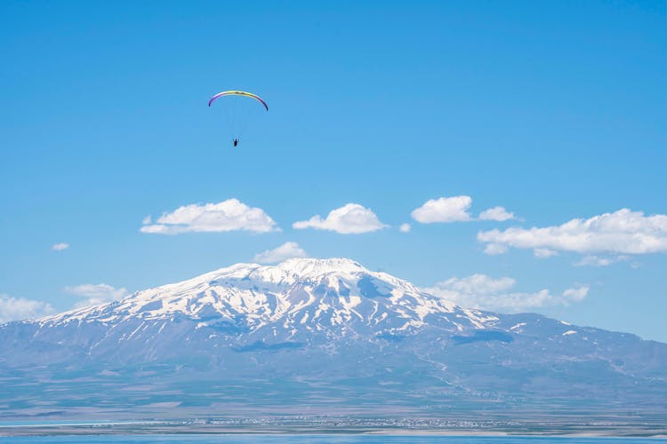 Person Paragliding Over The Sea Near Snow Covered Mountain