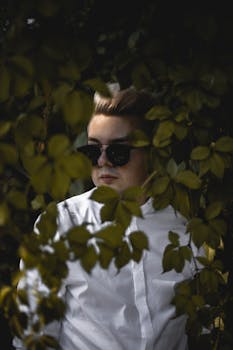 一名男子身穿白衬衫、戴着太阳镜，部分隐藏在户外郁郁葱葱的绿叶中