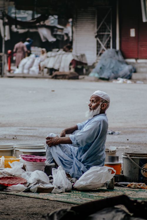 Free Senior man vending goods on a street, surrounded by bags and containers, in a busy urban environment. Stock Photo