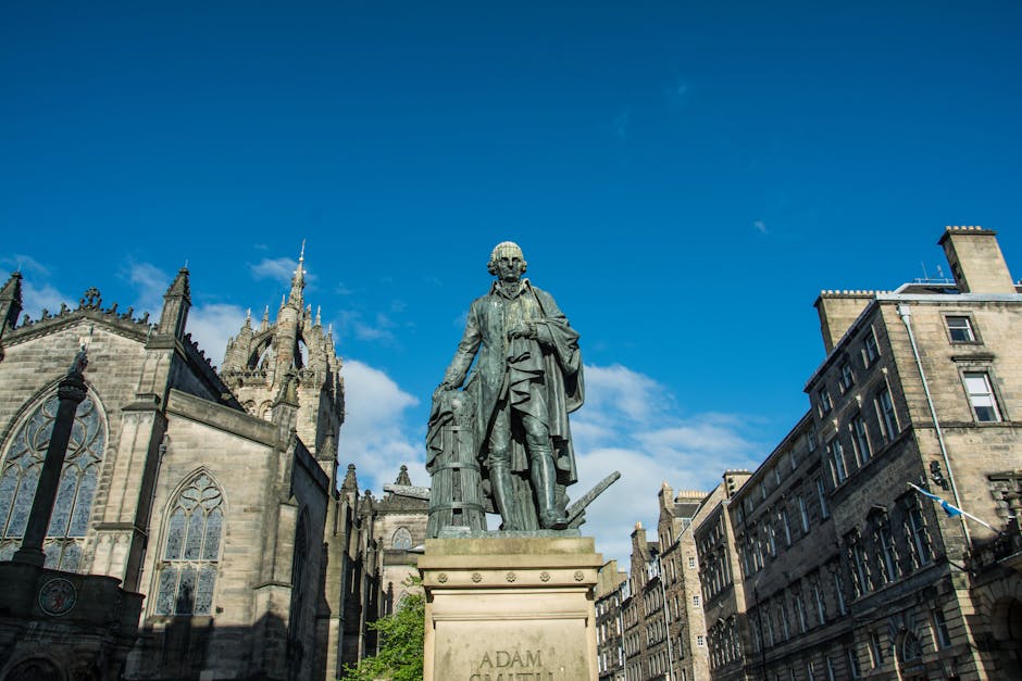 Statue of Adam Smith with historic buildings in Edinburgh, Scotland, under a blue sky.