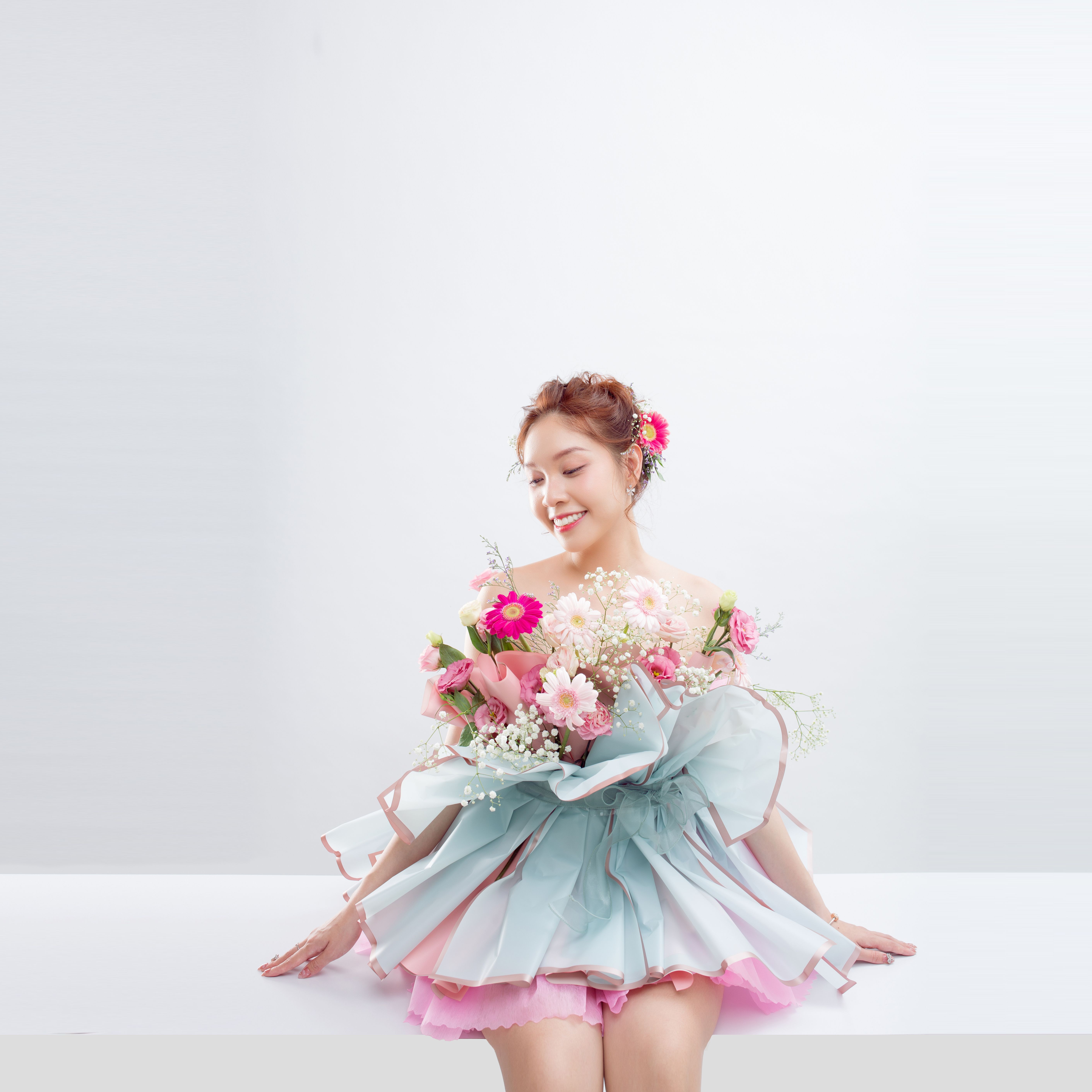Young woman in a pastel dress holding a floral bouquet, smiling in a studio setting.