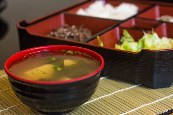 Black and red lacquer bowl of miso soup with tofu, wakame and scallions beside bento box