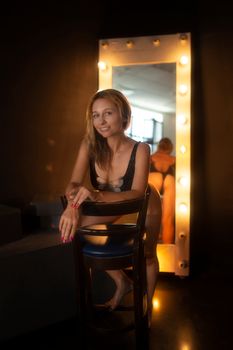 A woman sits confidently on a chair in lingerie with a lighted mirror backdrop.