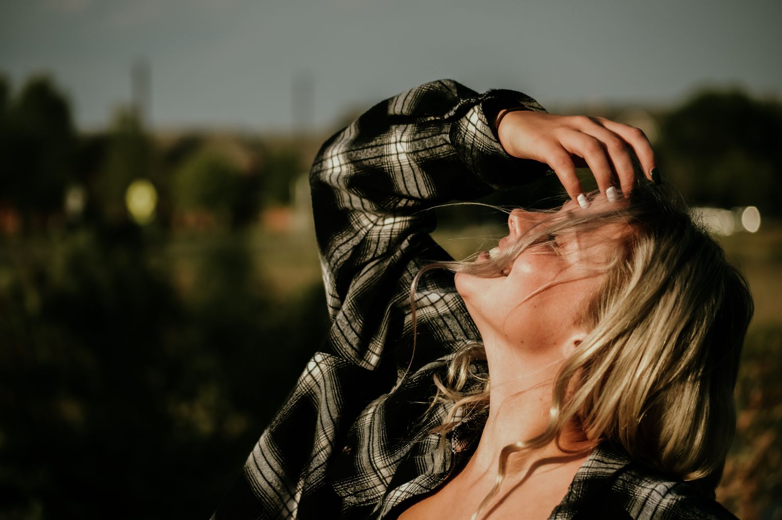 Blonde woman in a plaid shirt tilts her head back in golden sunlight, hand in her hair