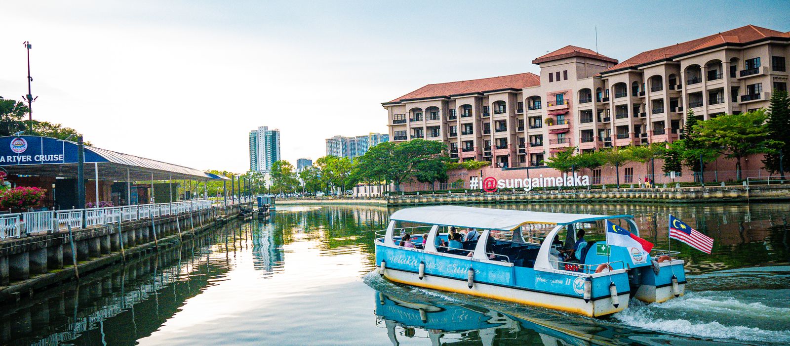 Colorful boat cruising on Melaka River with scenic view of buildings and blue sky.