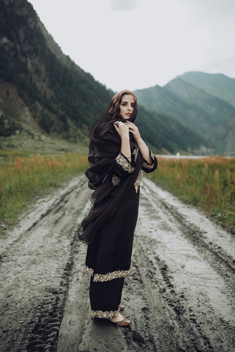 A Woman In Black Dress Standing On A Muddy Road