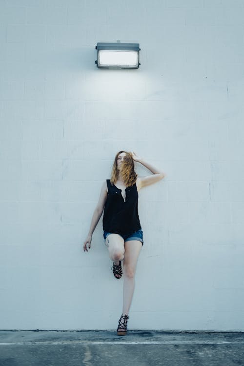 Woman in Black Tank Top and Shorts Leaning on White Wall