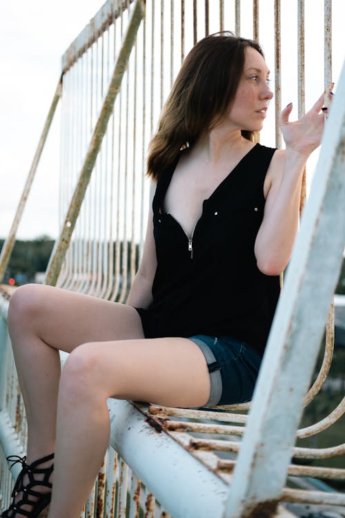 A Woman in Black Tank Top and Denim Shorts Sitting on Metal Railings