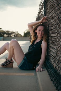 Stylish woman in denim shorts and wedges poses by a fence in natural light.