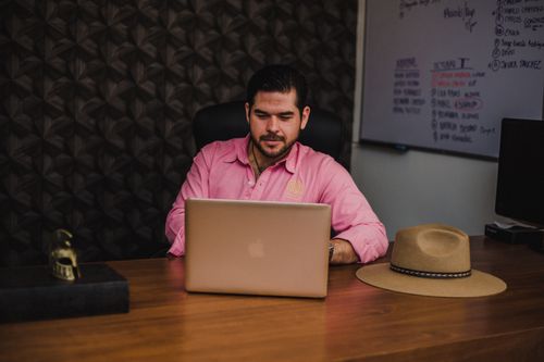 Free Man in pink shirt working on laptop at a wooden desk in office. Stock Photo