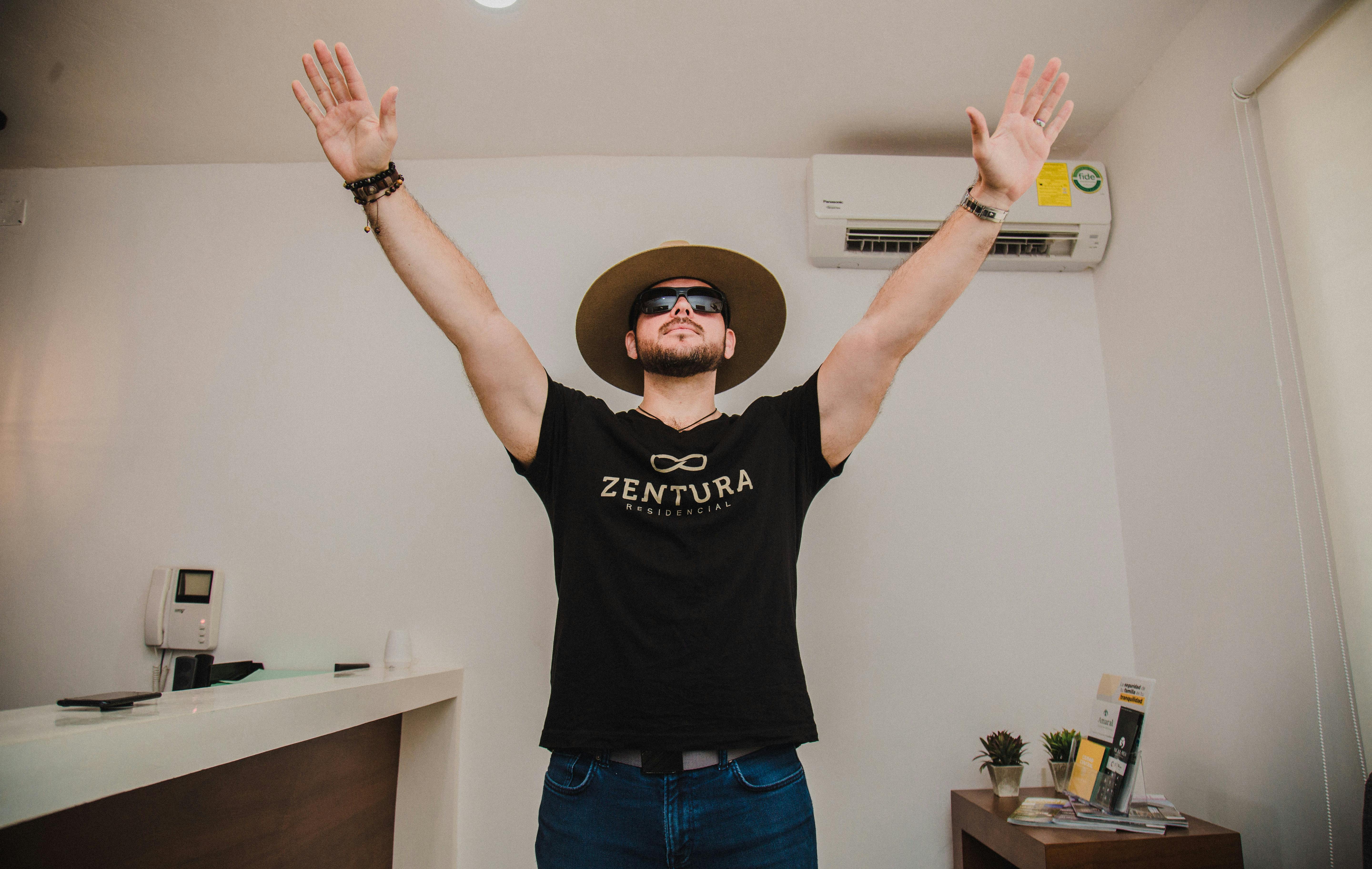 A young man stands indoors with arms raised, wearing a hat and sunglasses. Stylish and contemporary.