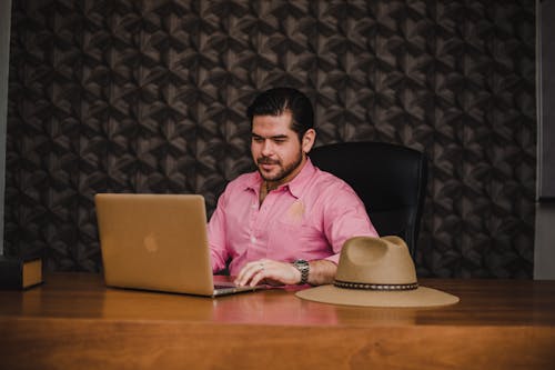 Free Man in Pink Dress Shirt Sitting on Black Chair in Front of Silver Macbook Stock Photo