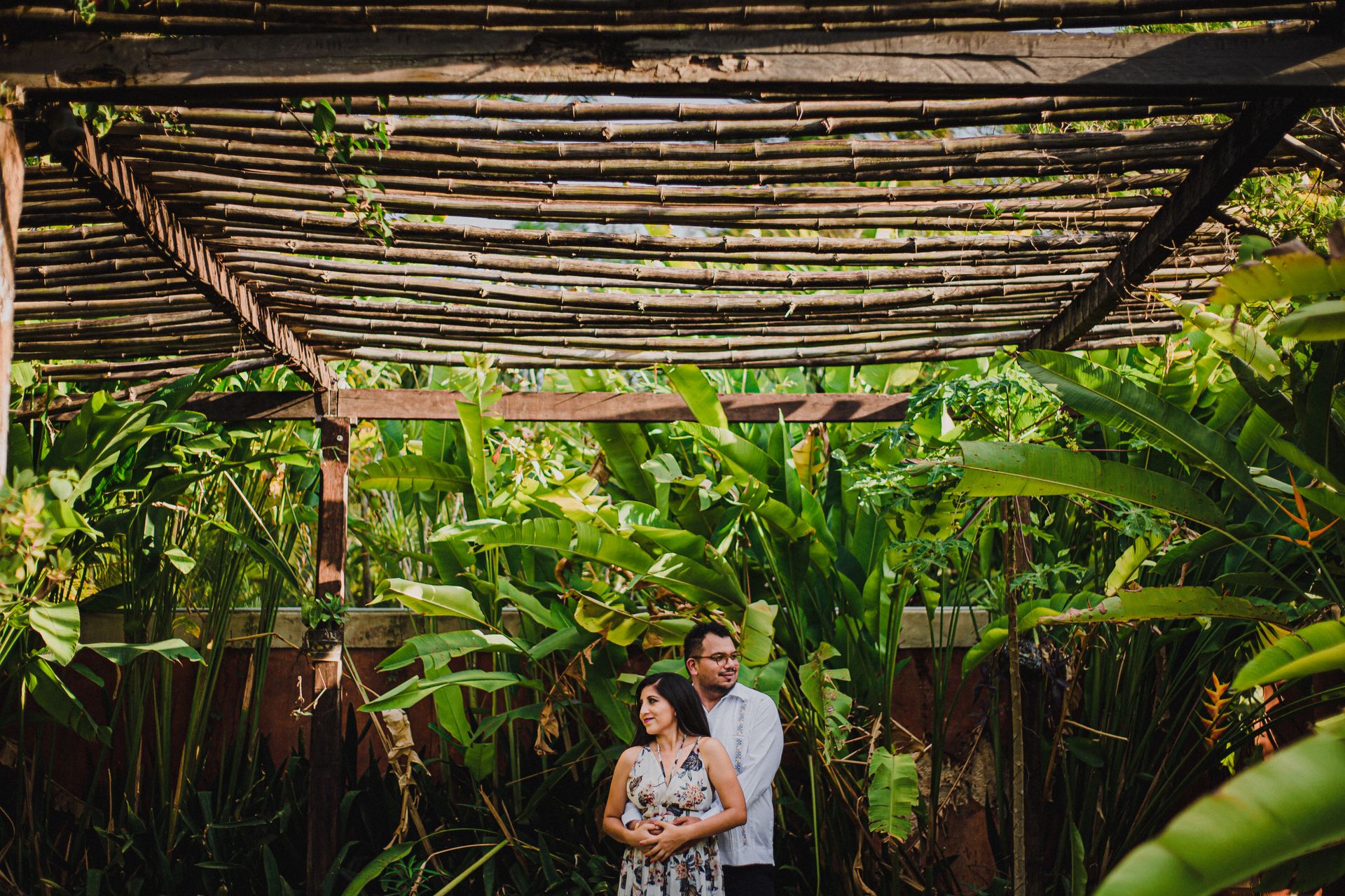 A couple embraces under a wooden pergola surrounded by lush green plants, depicting love and togetherness.