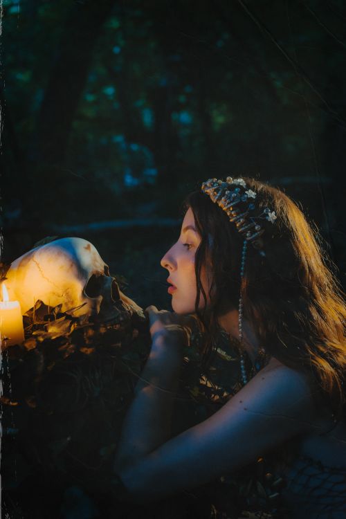 Free A mysterious woman with a wreath gazes at a skull by candlelight in a shadowy forest setting. Stock Photo