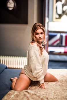 A woman wearing a knitted sweater poses elegantly on a bed indoors.