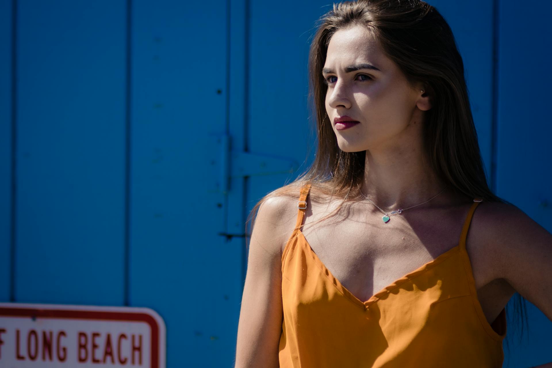 Stylish woman in orange top posing against a blue wall, Long Beach sign visible.