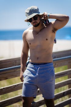Casual shirtless man with sunglasses and hat on a sunny beach boardwalk.