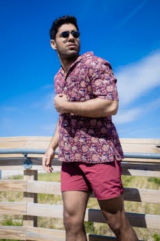 Adult man with sunglasses in floral shirt on wooden boardwalk against blue sky.